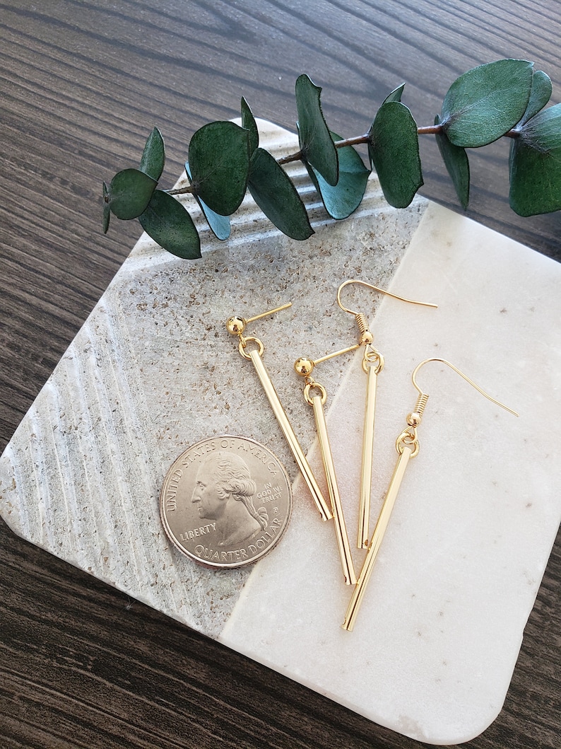 May include: Three pairs of gold bar earrings with a simple design. The earrings are on a white marble surface with a green plant branch.