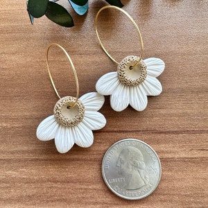 May include: A pair of gold hoop earrings with white and brown clay daisy flower charms. The earrings are shown on a wooden surface with a US quarter for scale.