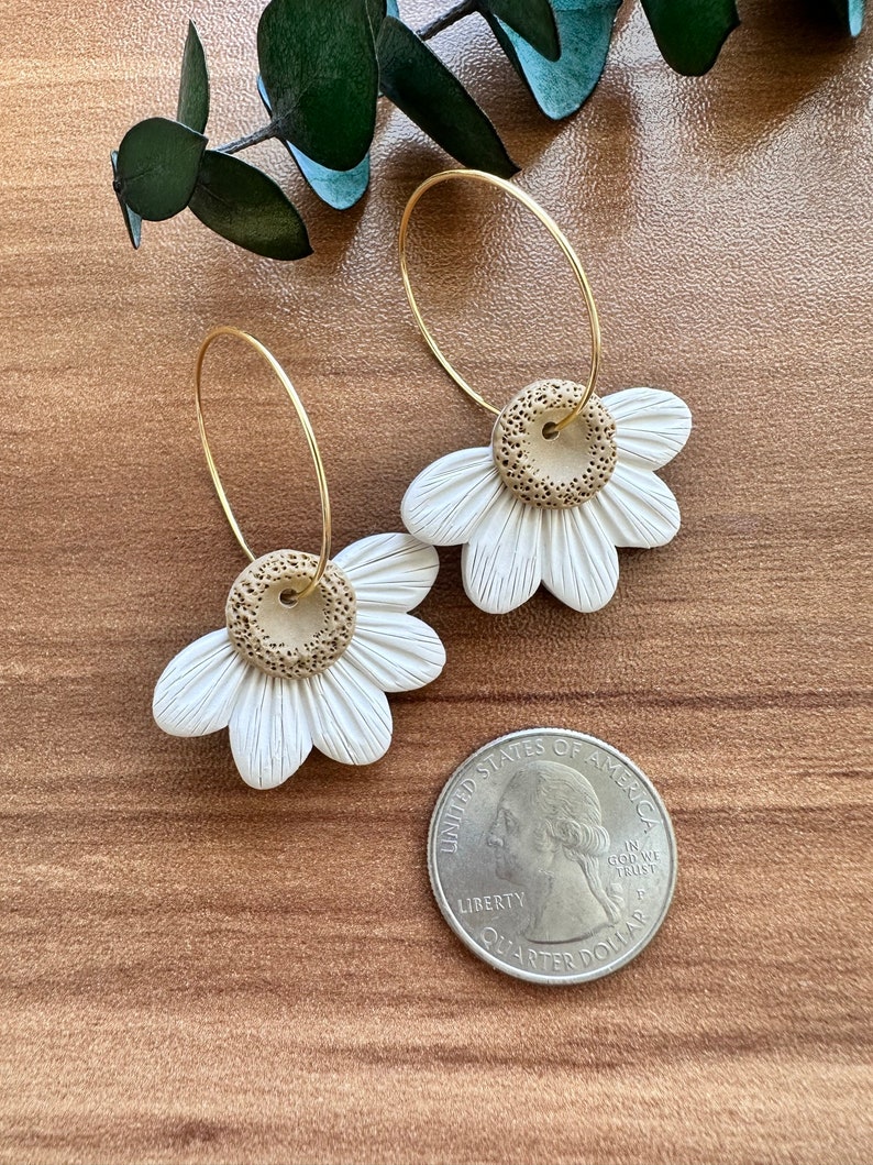 May include: A pair of gold hoop earrings with white and brown clay daisy flower charms. The earrings are shown on a wooden surface with a US quarter for scale.