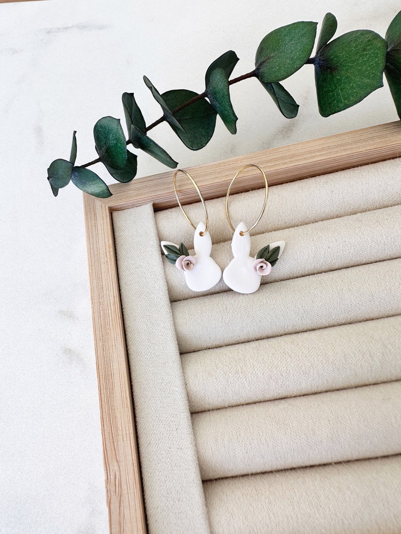 May include: A pair of gold hoop earrings with white bunny charms and pink flower accents. The earrings are displayed on a white velvet ring holder.