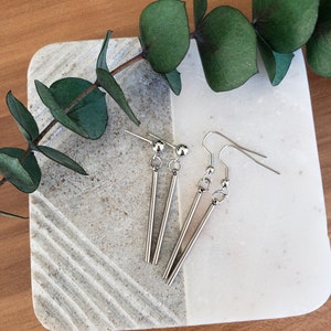 May include: A pair of silver dangle earrings with a long, thin bar pendant. The earrings have a simple, minimalist design.