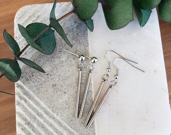 May include: A pair of silver dangle earrings with a long, thin bar pendant. The earrings have a simple, minimalist design.