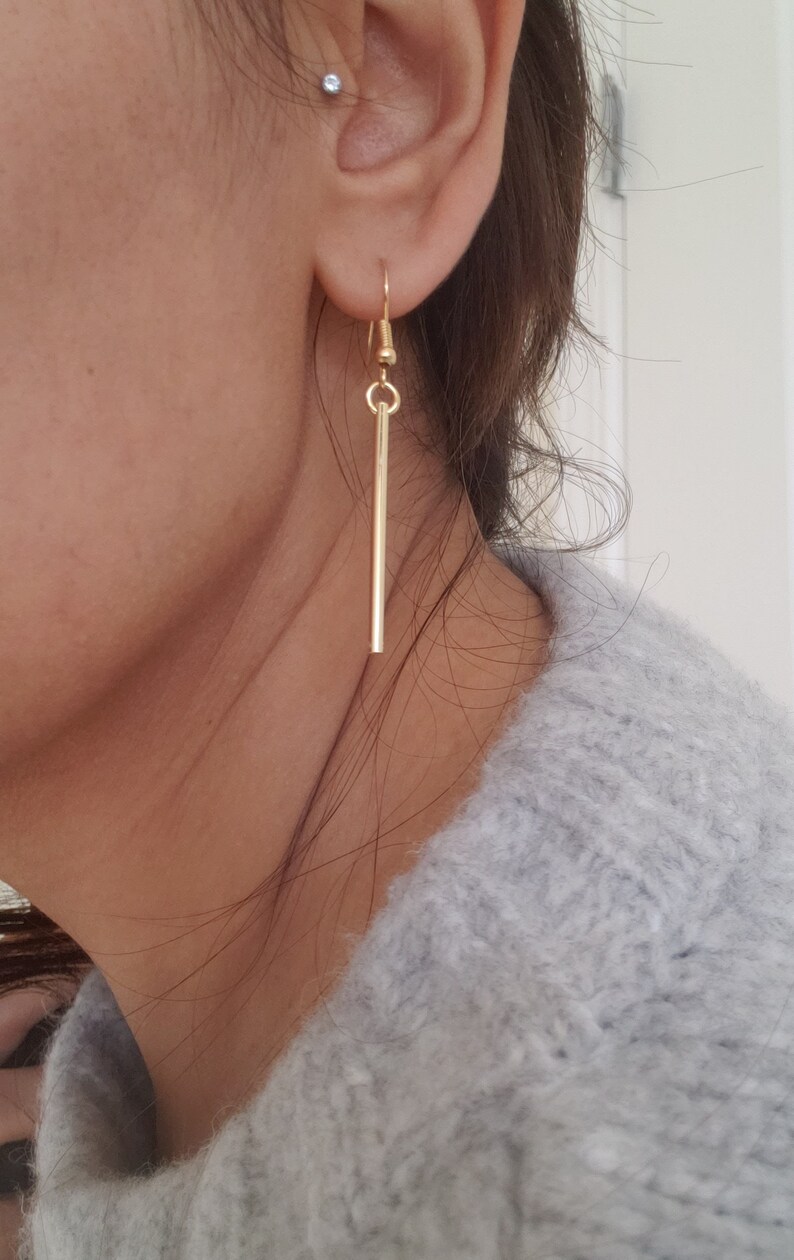 May include: Gold dangle earrings with a long, thin bar pendant.