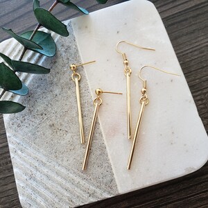 May include: A pair of gold-toned earrings with a long, straight bar design. The earrings are on a white marble surface.