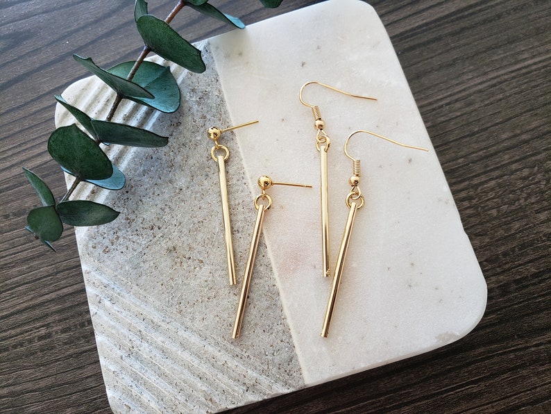 May include: A pair of gold-toned earrings with a long, straight bar design. The earrings are on a white marble surface.
