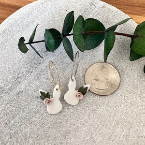 May include: A pair of white bunny-shaped earrings with pink flowers and green leaves. The earrings are on a white plate with a green plant in the background.