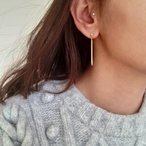 May include: A pair of gold bar earrings with a small ball at the top. The earrings are hanging from the earlobe.