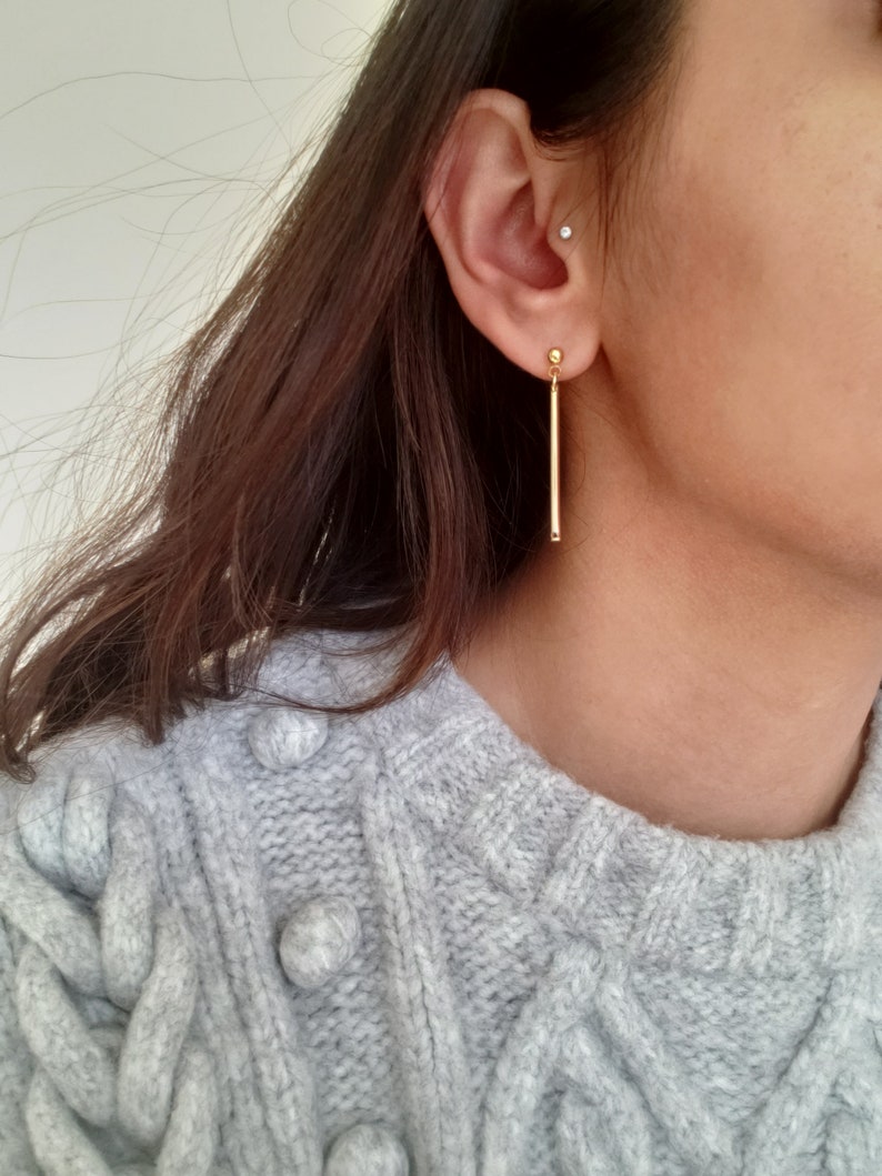 May include: A pair of gold bar earrings with a small ball at the top. The earrings are hanging from the earlobe.