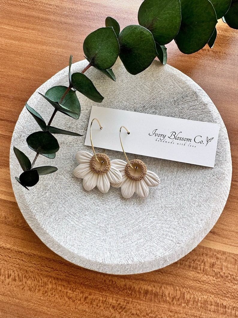 May include: A pair of white daisy earrings with gold hoops and brown centers. The earrings are displayed on a white plate with a card that reads "Ivory Blossom Co. Handmade with love."