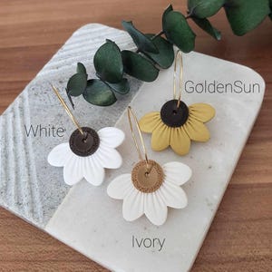 May include: Three pairs of flower-shaped earrings with gold hoops. The earrings are in three colors: white, ivory, and yellow. The text "White", "Ivory", and "GoldenSun" is written below each pair of earrings.