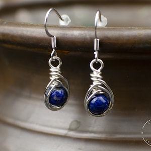 May include: A pair of silver wire wrapped earrings with blue lapis lazuli stones. The earrings are hanging from a silver hook.
