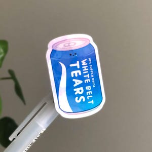 May include: A sticker in the shape of a blue and white can with the words "WHITE BELT TEARS" and "ION SUPPLY DRINK" printed on it. The can has a pink top and a small smiling face.