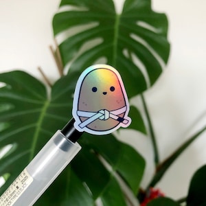 May include: A holographic sticker of a cartoon potato wearing a white karate belt. The potato has a brown body and a happy face.