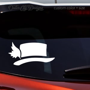 May include: White vinyl decal of a top hat with a wing on the side. The decal is on a car window. The text "Outer City Designs" and "custom color + size" is visible in the background.