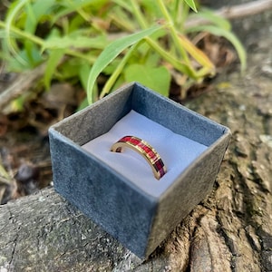 May include: A gold-colored ring with a band of red and burgundy stones is displayed inside a small, dark gray box. The ring is set against a white background. The box is resting on a weathered, textured surface with green foliage in the background.