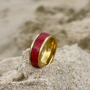 Puede incluir: Un anillo dorado con una llamativa incrustación roja. El anillo se muestra sobre arena, resaltando su acabado pulido y el color intenso de la incrustación. El diseño del anillo es simple pero elegante, adecuado para el uso diario.