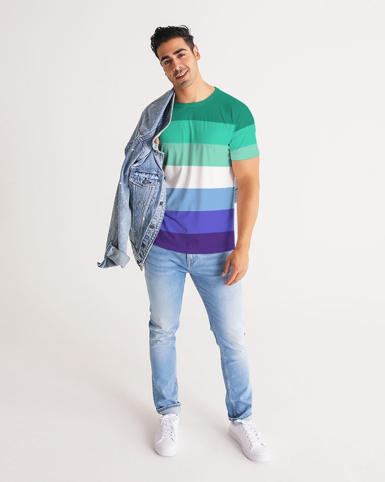 Puede incluir: Una persona lleva una camiseta a rayas verde, blanca, azul y morada con una chaqueta vaquera sobre los hombros. Lleva pantalones vaqueros azules claros y zapatillas blancas.