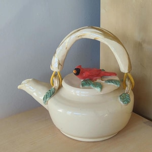 May include: A white ceramic teapot with a cardinal bird on the lid and a brown and white handle. The teapot has green leaves around the base and lid.
