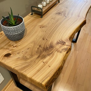 May include: A live edge wood console table with a natural wood finish and black metal legs. The table has a unique, organic shape and is decorated with a small potted succulent and a metal candle holder with five white candles.