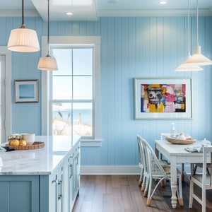 Puede incluir: Una cocina azul claro con una isla de encimera de m&aacute;rmol blanco, una ventana con vistas al oc&eacute;ano y una mesa de comedor con sillas blancas. La pared est&aacute; pintada de azul claro y tiene paneles de madera blanca. Hay una pintura abstracta colorida en la pared.