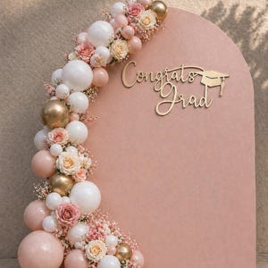 May include: A pink arched backdrop with the words "Congrats Grad" in gold script, adorned with a balloon garland of white, gold, and pink balloons, and roses. A graduation cap is next to the text.