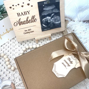 May include: A wooden baby announcement frame with the name "Anabelle" and a sonogram image. A gift box with a gold ribbon and a tag that says "Baby coming soon..." is also present. A card that says "Congratulations" is also visible.