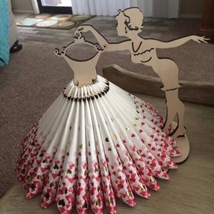 May include: A wooden napkin holder shaped like a woman holding a dress. The dress is made of white paper napkins with red and gold hearts printed on them.