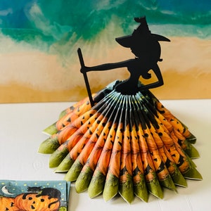 May include: A black silhouette of a witch holding a broom sits atop a fan of orange and green napkins with a pumpkin design. The napkins are folded into a fan shape, creating a festive Halloween centerpiece.
