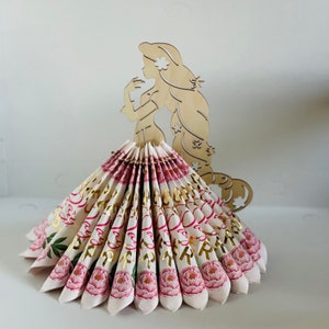 May include: A wooden cutout of a princess with long flowing hair, holding a napkin holder shaped like a pink and white ruffled skirt with gold lettering and floral accents.