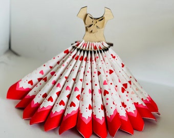 Könnte beinhalten: Ein weißer und roter Serviettenhalter in Form eines Kleides mit einer Holzschleife am Dekolleté. Das Kleid ist mit rosa und roten Herzen und weißen Punkten verziert.