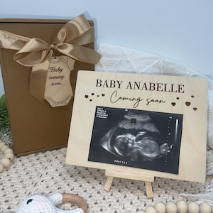 May include: A wooden sign with the text "BABY ANABELLE Coming soon" and a sonogram image. The sign is on a small easel. A brown gift box with a gold ribbon and a wooden tag that says "Baby coming soon..." is next to the sign.
