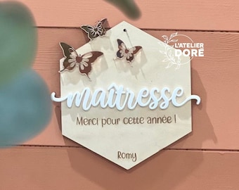 May include: A wooden hexagonal sign with the words "maîtresse" and "Merci pour cette année!" in white script. The sign is decorated with three butterfly cutouts and the text "L'ATELIER DORE" in a circle. The sign is on a pink wooden background.