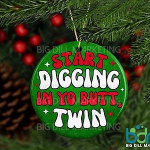 May include: A round, green Christmas ornament with the text "START DIGGING IN YO BUTT, TWIN" in white and red lettering. The ornament is hanging from a gold string against a backdrop of pine needles and red berries.