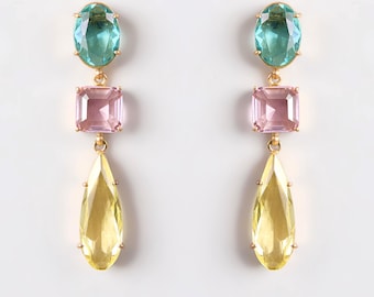 May include: A pair of gold-toned earrings with three colorful gemstone shapes. Each earring features an oval teal stone, a square pink stone, and a teardrop-shaped yellow stone. The earrings are displayed against a white background.