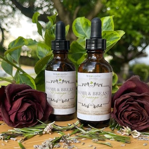 以下が含まれることがあります： 黒いスポイト付きの2つの琥珀色のガラス瓶に入ったマッサージオイル。「Womb & Breast Massage Oil」とラベルされています。ボトルは、乾燥したバーガンディ色のバラ、ローズマリーの小枝、ラベンダーのつぼみで囲まれています。木製の表面に置かれ、背景には緑の葉があります。