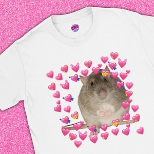 May include: White t-shirt featuring a graphic of a brown rat surrounded by pink hearts with blue arrows. The shirt has a crew neck and a small logo on the upper right. The text "CryCouture x YesBabeStudio Collection" is at the bottom.