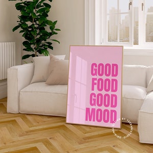 May include: A pink framed print with the words "GOOD FOOD GOOD MOOD" in bold pink letters. The print is in a modern living room with a white sofa, cushions, and a large window overlooking a cityscape.