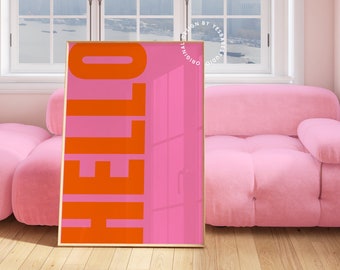 May include: A pink and orange poster with the word "HELLO" in large, bold, orange letters. The poster is framed in gold and is sitting on a wooden floor in front of a pink sofa.