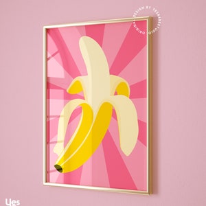 May include: A framed print of a yellow banana on a pink background with a sunburst pattern. The print is in a gold frame.