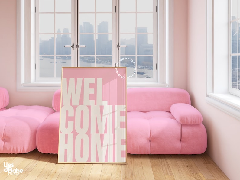 May include: A pink and white poster with the text "WELCOME HOME" in a bold, white font. The poster is framed in gold and is leaning against a pink sofa.
