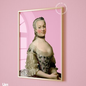May include: A framed portrait of a woman in a white and pink dress with a lace collar. The woman has a light complexion and is wearing a white wig. The background is a light pink color. The frame is gold.