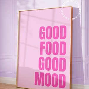 May include: A pink poster with the words "GOOD FOOD GOOD MOOD" in bold, bright pink letters. The poster is in a gold frame and leans against a light purple wall. The words "Yes Babe Studio" are in the bottom left corner.