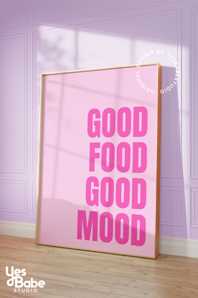 May include: A pink poster with the words "GOOD FOOD GOOD MOOD" in bold, bright pink letters. The poster is in a gold frame and leans against a light purple wall. The words "Yes Babe Studio" are in the bottom left corner.