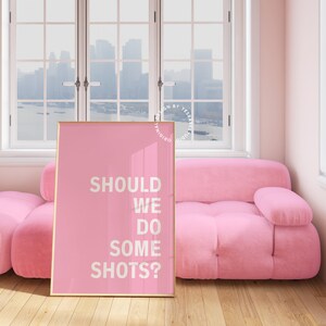 May include: Pink framed poster with the text "SHOULD WE DO SOME SHOTS?" on a pink background. The poster is leaning against a pink sofa in a room with windows.