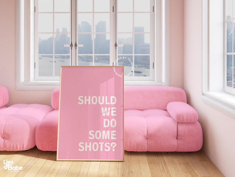 May include: Pink framed poster with the text "SHOULD WE DO SOME SHOTS?" on a pink background. The poster is leaning against a pink sofa in a room with windows.