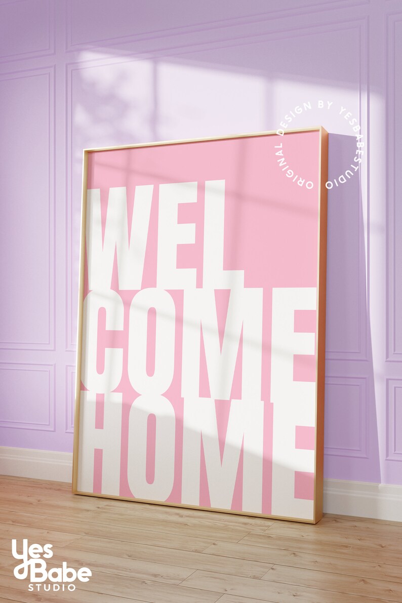 May include: A pink and white framed print with the text "WELCOME HOME" in bold white letters. The print is in a gold frame and is hanging on a light purple wall.