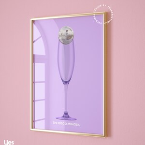 May include: A framed print with a purple background featuring a champagne flute with a disco ball on top. The text "THE DISCO MIMOSA" is printed below the glass. The frame is gold.