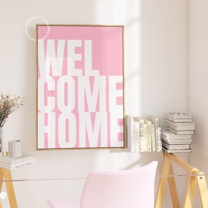 May include: A pink canvas print with a white inscription that reads "WELCOME HOME" in a bold, blocky font. The print is framed in a thin, gold frame and is hanging on a white wall.