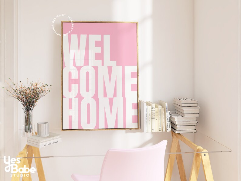 May include: A pink canvas print with a white inscription that reads "WELCOME HOME" in a bold, blocky font. The print is framed in a thin, gold frame and is hanging on a white wall.