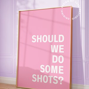 May include: A pink framed poster with the text "Should we do some shots?" in white lettering. The poster is hanging on a light purple wall.